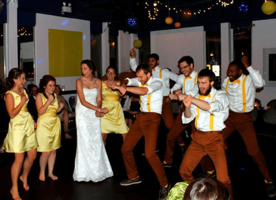 A group of people dance at a wedding. On the left, a bride stands in a white dress with her bridesmaids in short yellow dresses, and they laugh. To their right, young men in brown pants and white collared shirts with yellow suspenders dance with knees slightly bent and arms crossed out in front of their torsos.